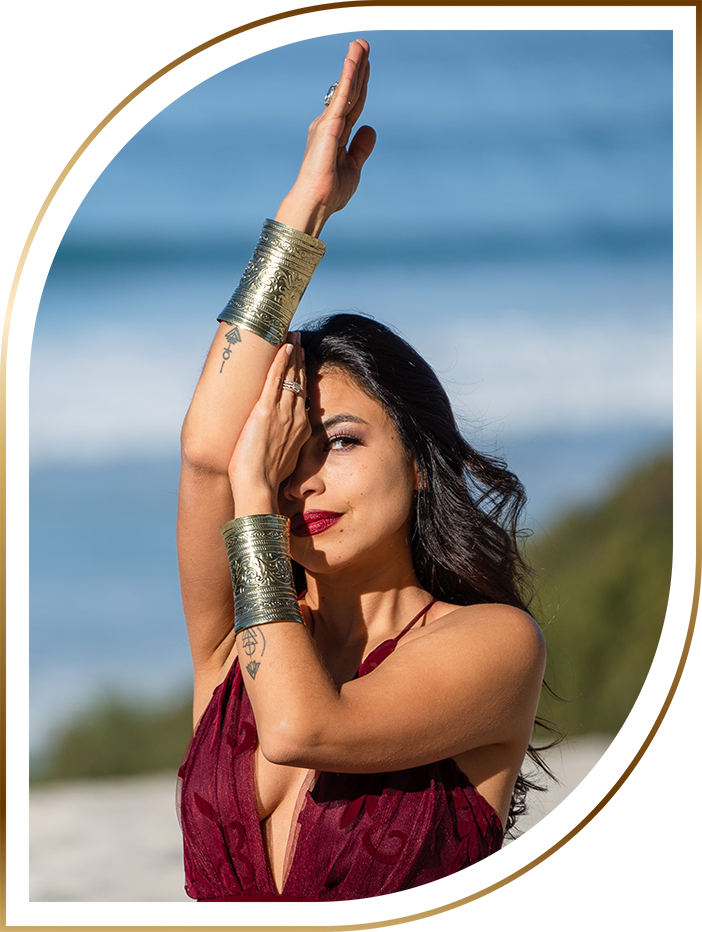 Safira Vinci posing outdoors with arms raised, wearing gold cuff bracelets and a burgundy dress against a blue sky