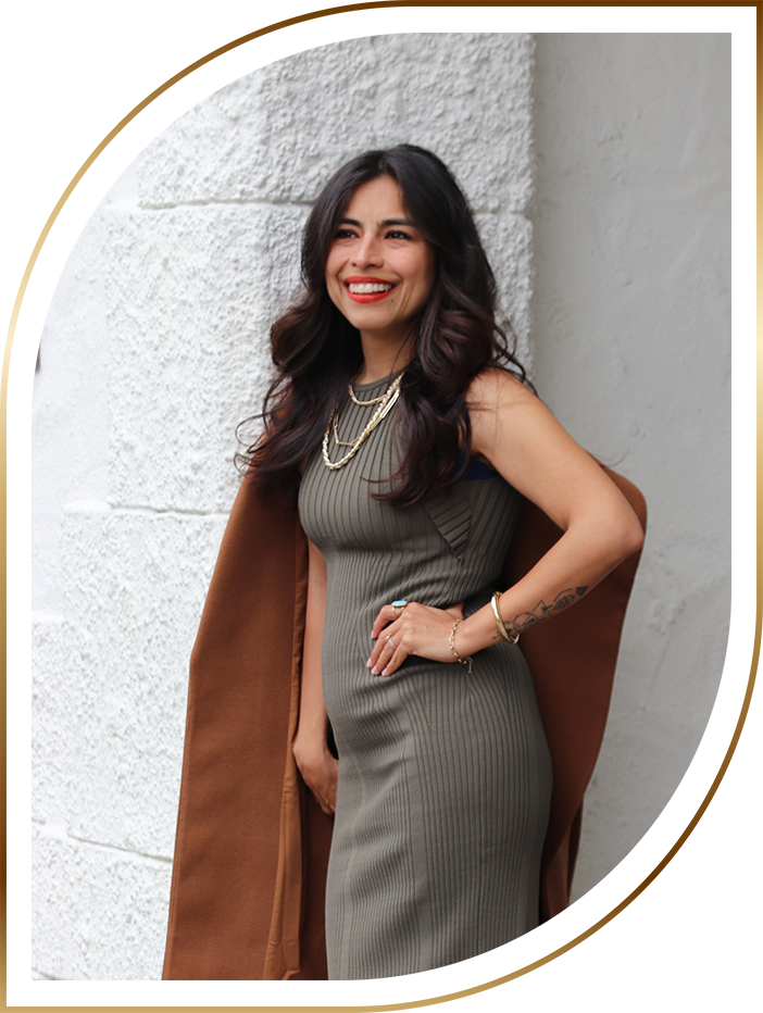 Safira Vinci smiling and standing against a white wall, wearing a fitted dress with a brown coat draped over her shoulders